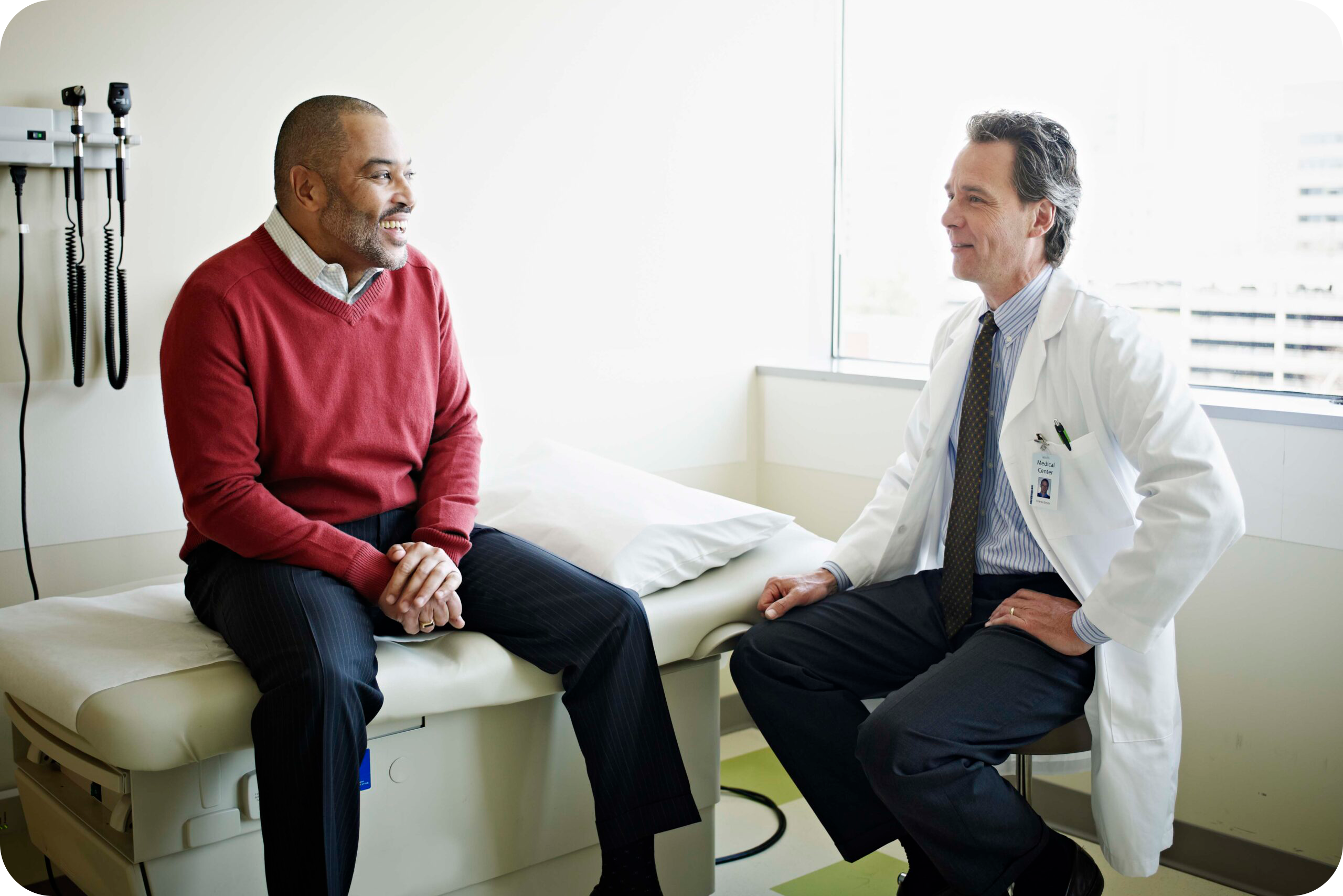 Older Black male patient sits on an exam table as he talks to a middle age White male doctor.
